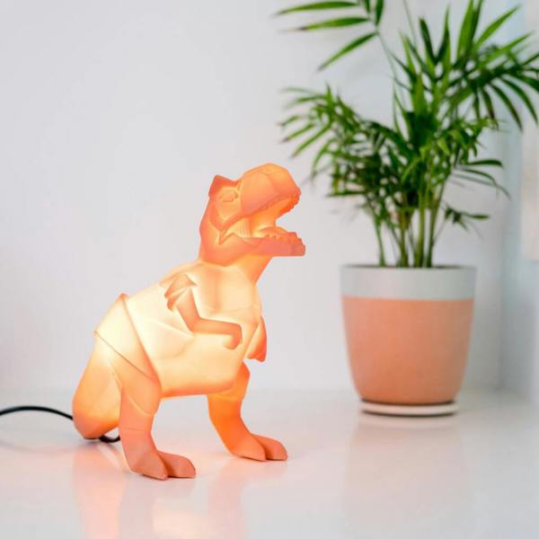 Glowing orange origami T-Rex dinosaur lamp sitting on a white desk next to a potted houseplant