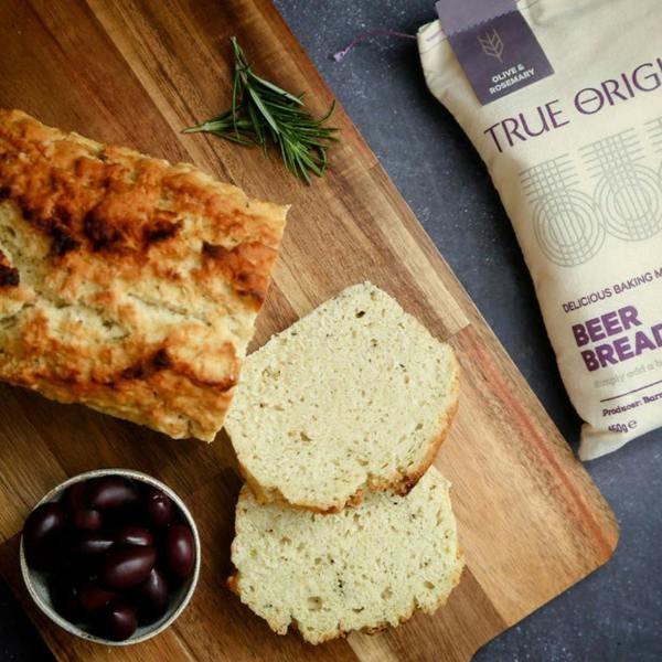 A freshly baked and sliced loaf of Olive and Rosemary beer bread on a wooden chopping board, next to a fabric sack of mix and a small bowl of dark olives.