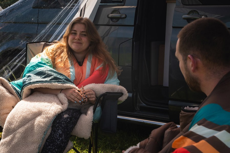 Insulated Outdoor Blanko OLPRO worn by a young lady and a young man, wrapped in the Blanko blankets whilst sat chatting, one wearing the insulated tonal tree tops with sherpa fleece and one wearing the vivid spruce. imageset:Tonal Tree Tops