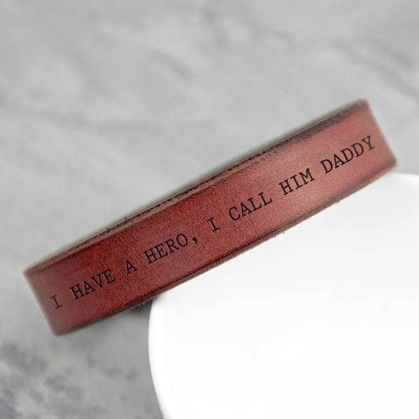 Close-up of a thick, personalised brown leather cuff bracelet with a custom engraved message, 'I HAVE A HERO, I CALL HIM DADDY', resting on a white surface.