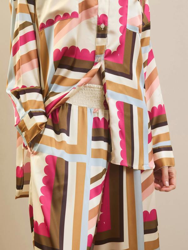 A close-up of a person wearing a matching long-sleeve button-front pyjama top and skirt set with a bold geometric pattern in beige, brown, pink, light blue, and white.