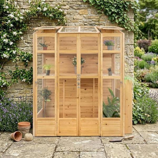 Victorian greenhouse in natural wood, featuring glass panels, shelves for plants, and a rustic stone wall backdrop with greenery