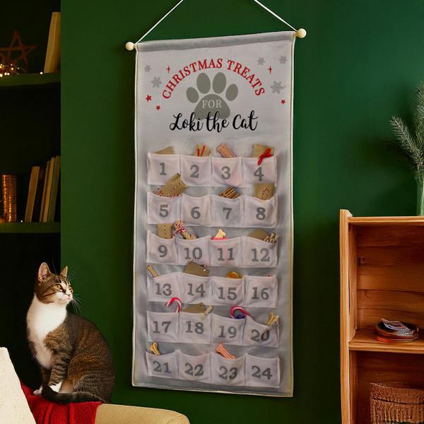 A tabby cat sitting on the edge of a couch next to a personalised grey fabric advent calendar reading "Loki the Cat" on a green wall.