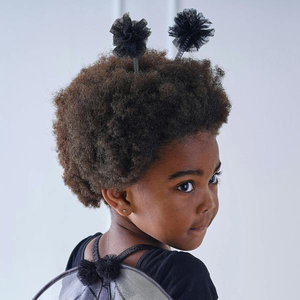 Over-the-shoulder portrait of a young girl with an afro wearing a bumble bee headband with black tulle pom pom antennae.
