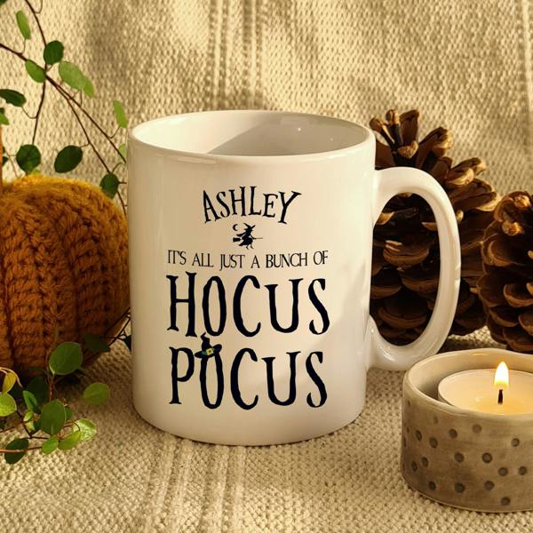 White ceramic mug reading Ashley It's All Just A Bunch Of Hocus Pocus next to a candle and pumpkin
