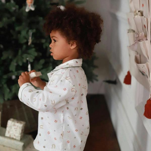 A side profile view of a young child wearing white organic cotton flannel Christmas pyjamas with a Nutcracker print, holding a small wrapped package by a Christmas tree.