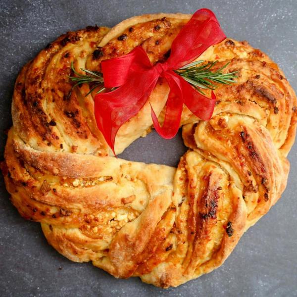 A freshly baked savoury beer bread loaf shaped into a decorative festive wreath, garnished with fresh rosemary sprigs and a red ribbon bow on a dark slate surface.
