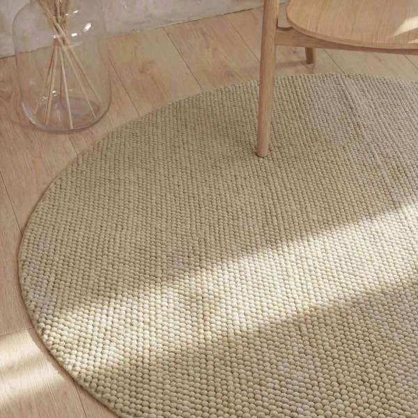 Round woven rug on a wooden floor with a glass jar and wooden chair.