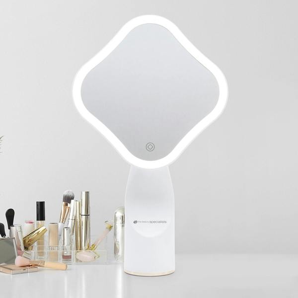 The Rio Illuminated Full Size Beauty Mirror with a white frame sits on a white table, surrounded by neatly arranged makeup brushes, cosmetics, and beauty products against a plain light gray wall.