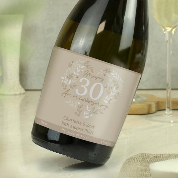 A tilted bottle of personalised 30th Pearl Wedding Anniversary Prosecco on a table next to a champagne glass, with the custom label from Abode Home & Gifts showing text reading Charlotte and Jack 18th August 2024.