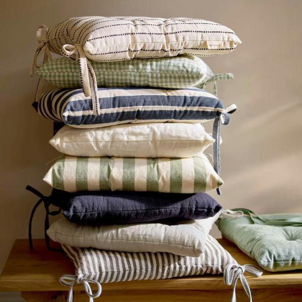 A high stack of various linen seat pads in solid colours and stripes on a wooden bench.