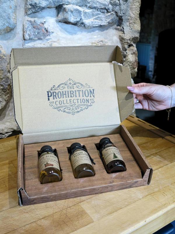 A hand opens a cardboard box labeled 'The Prohibition Collection' resting on a wooden table, revealing three small amber glass bottles in a wooden tray.