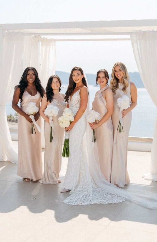 Bridesmaids in elegant dresses with floral bouquets by a window with a view of water.