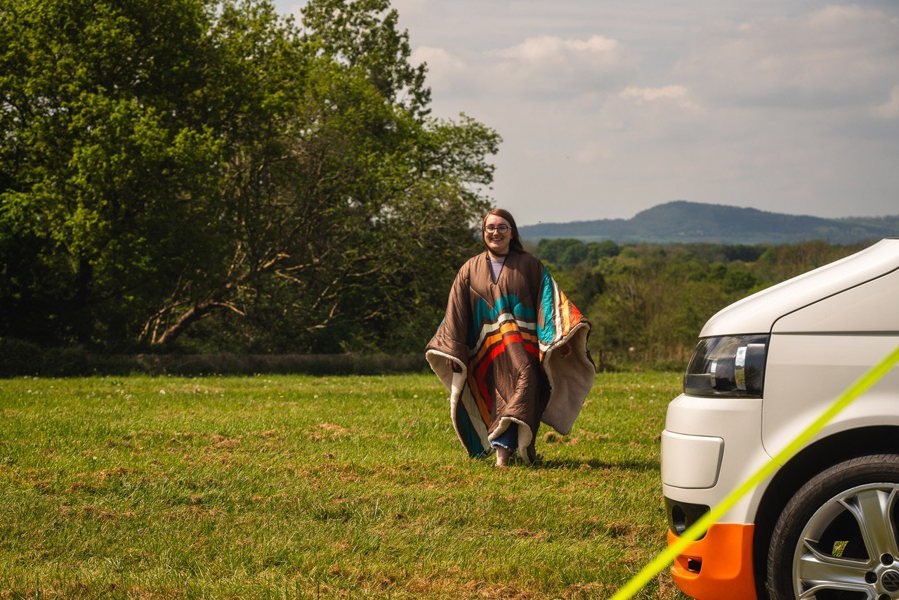 a young lady walking towards a camper van wearing the Vivid Spruce Insulated Outdoor Blanko Blanket as a poncho. imageset:Vivid Spruce