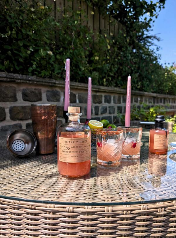 A bottle of guava picante by prohibition cocktails and two matching glasses with pink straws sit on a wicker table outdoors, alongside a copper shaker and lime slices, with a stone wall and greenery in the background.