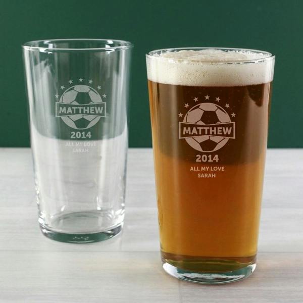 Two custom-engraved pint glasses set against a dark green painted wall background; one glass is empty and the other is filled with foaming amber beer, both featuring a football graphic etched with 'MATTHEW 2014 ALL MY LOVE SARAH'.