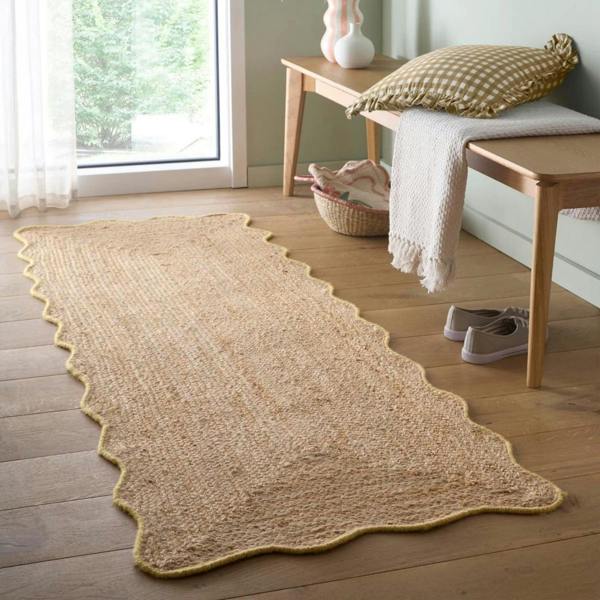 Beige woven rug on a wooden floor with a bench and pillows in the background.