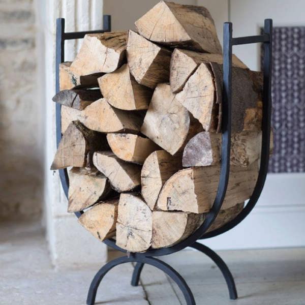 A high-sided black wrought iron log holder filled with neatly stacked split firewood logs, standing on a light stone hearth next to a rustic feature wall.