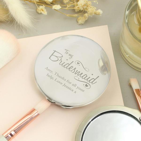 Front view of the engraved nickel-plated bridesmaid compact mirror sitting straight next to its black fabric pouch on a white background.