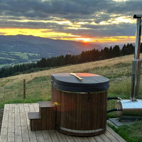 Wood-fired hot tub on a wooden platform with a scenic sunset over mountains and fields.