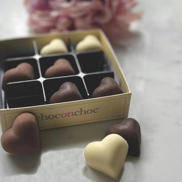 An open square gift box containing a variety of white, milk, and dark chocolate hearts on a white marble tabletop surface with soft pink flowers blurred in the background.