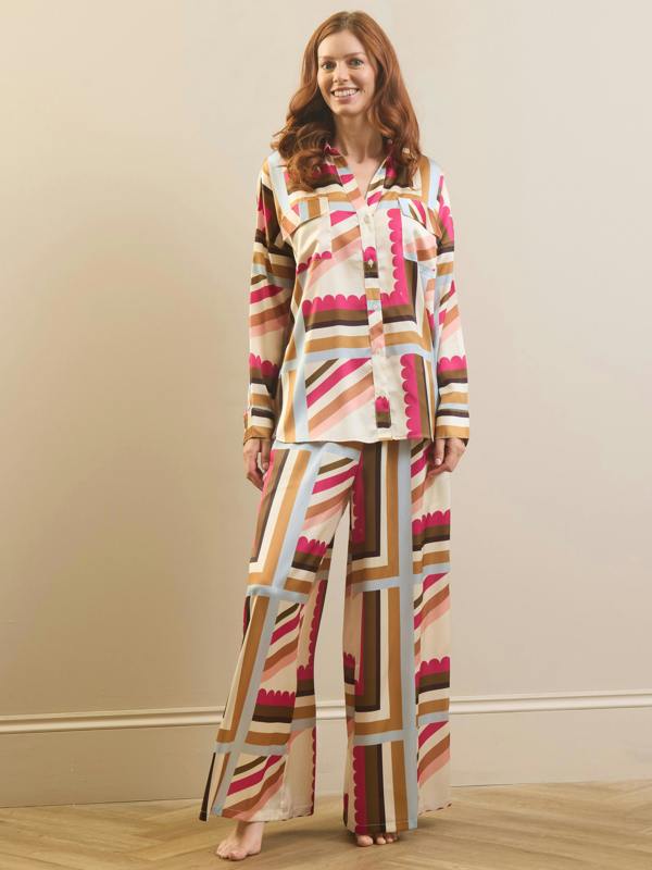 A woman modeling a matching two-piece pajama set with a retro geometric stripe pattern in pink, brown, beige, and light blue on a cream background, standing against a beige wall.