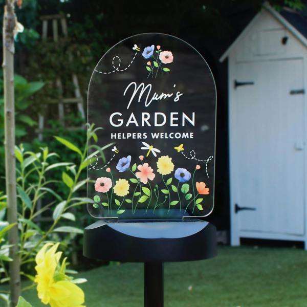 An arched clear acrylic garden solar light stake glows brightly outdoors on a black pedestal base. The sign is custom printed with colourful wildflowers, bumblebees, and a dragonfly around the text 'Mum's GARDEN, HELPERS WELCOME'. A white wooden garden shed and green foliage are softly blurred in the background.