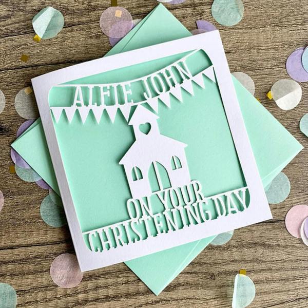 Personalised papercut christening card with the name Alfie John on bunting above a church against a mint green paper insert