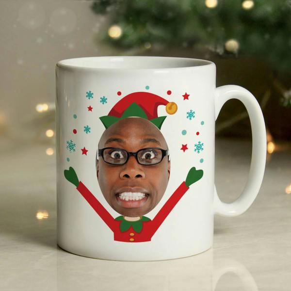 A white ceramic mug featuring a custom face photo inside a red and green Christmas elf illustration, set against a blurred background of festive lights.