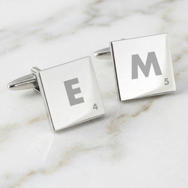 A pair of silver Scrabble tile cufflinks resting on a white marble surface, displaying the engravings 'E 4' and 'M 5'.
