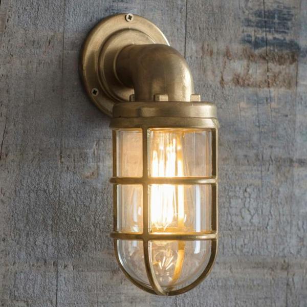Nautical raw cast brass down light with an industrial cage design and lit filament bulb, mounted on a weathered wood wall.