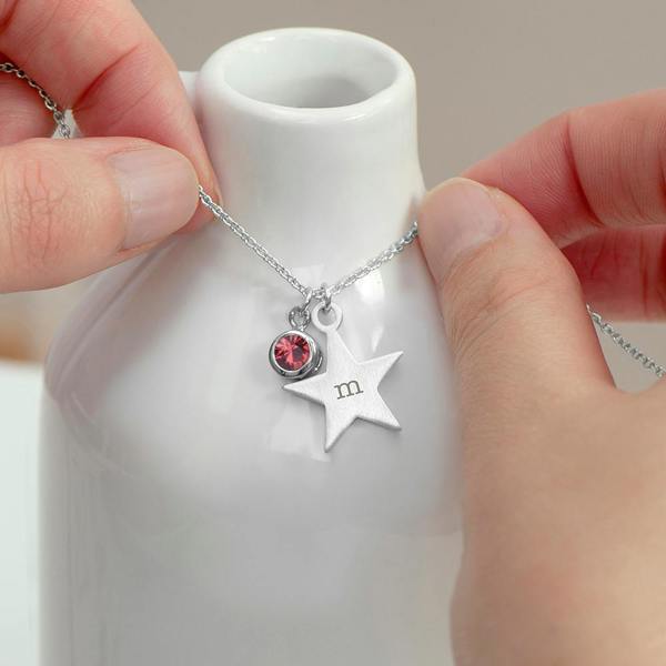 Two hands holding a delicate silver chain necklace around the neck of a glossy white ceramic vase, featuring a small brushed-silver star pendant engraved with a lowercase letter 'm' alongside a round pink crystal birthstone charm.