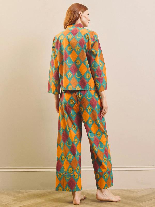 A woman stands barefoot indoors wearing a matching two-piece pyjama set with a colorful harlequin diamond pattern in orange, teal, and burgundy. The set includes a long-sleeve top and wide-leg pants, shown from the back against a plain beige wall.