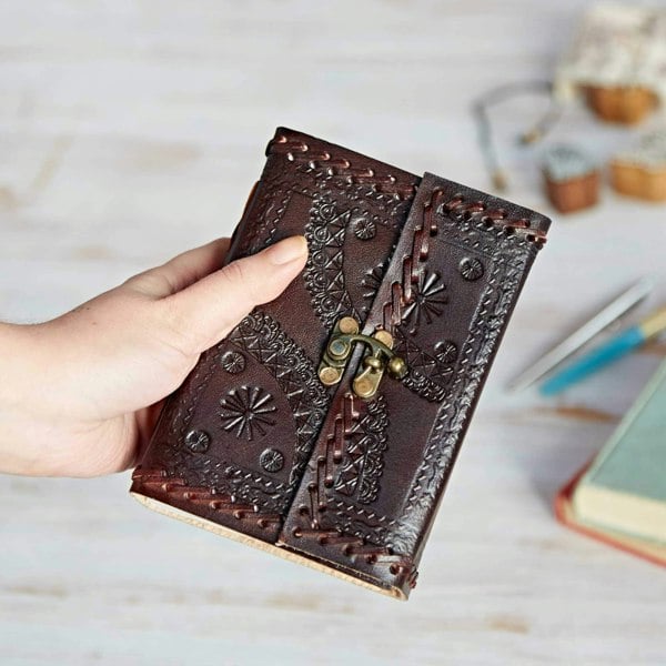 The Indra Medium Embossed & Stitched Leather Journal, with its intricate designs and sturdy metal clasp, is held against a blurred backdrop of books and a pen.