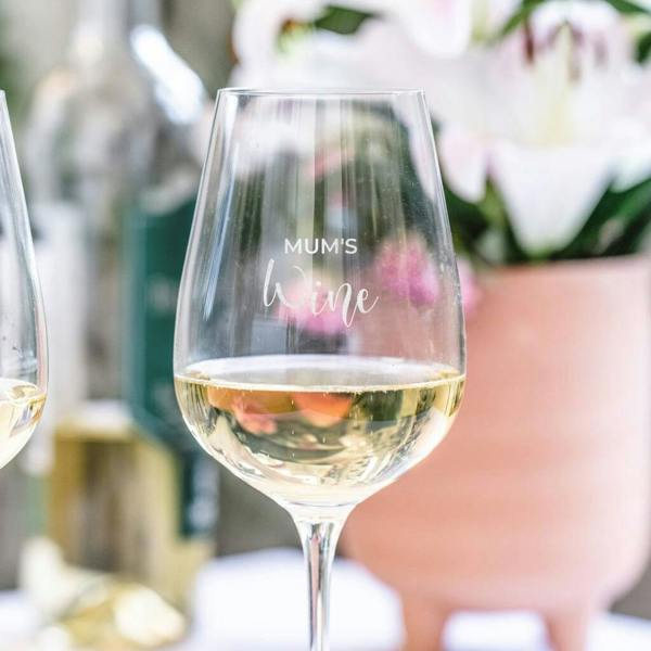 A clear stemmed wine glass filled with white wine, engraved with Mum's Wine, set outdoors against a soft background of pink flowers and wine bottles.