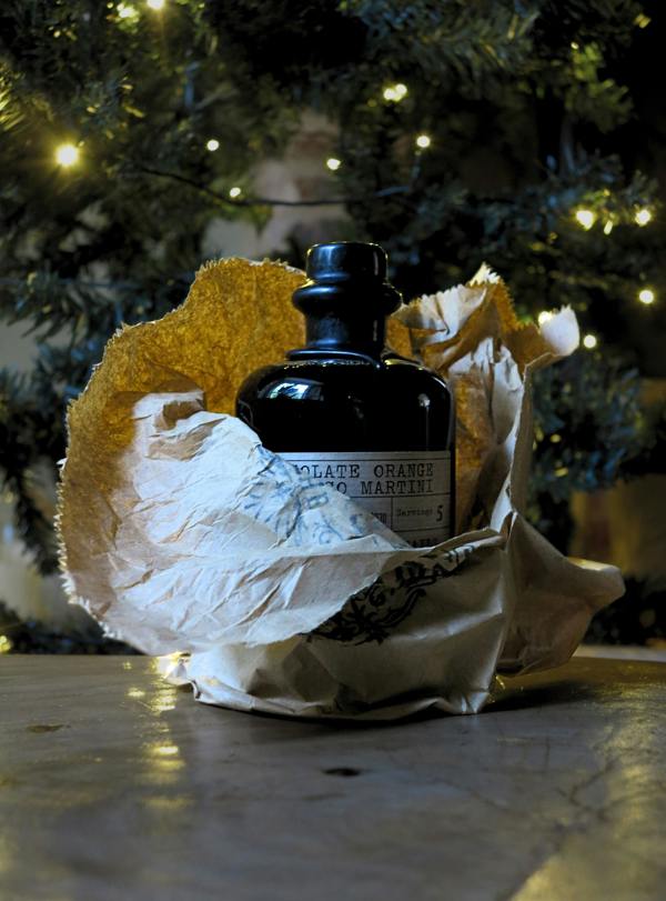 A dark glass bottle with a black stopper and a partially visible label reading 'chocolate orange espresso martini' sits wrapped in crumpled white paper with a gold textured accent, placed on a tabletop in front of a softly blurred Christmas tree with warm lights.