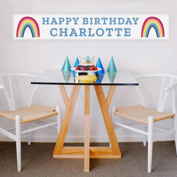 A white vinyl party banner printed with two colourful rainbows flanking the blue text HAPPY BIRTHDAY CHARLOTTE, hanging on a wall above a small wooden table styled with a birthday cake and party hats.