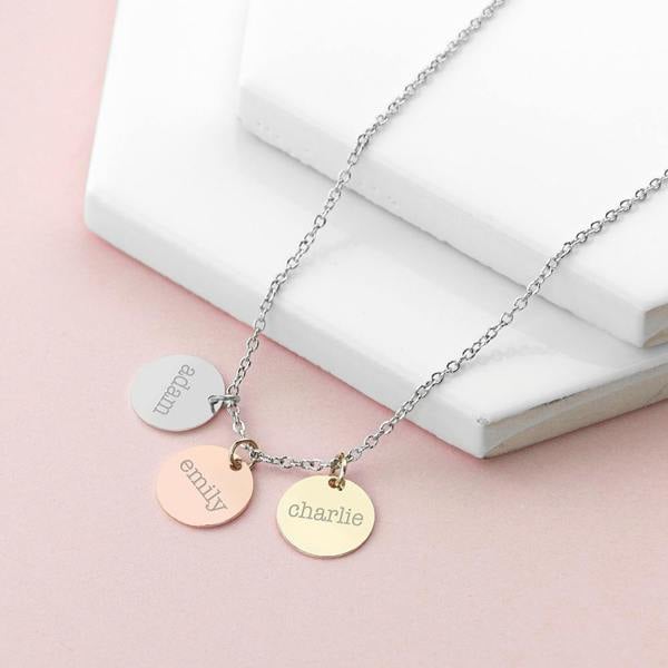 A personalised mixed-metal three disc necklace featuring matte silver, rose gold, and yellow gold charms engraved with the names 'adam', 'emily', and 'charlie' on a pink and white background.
