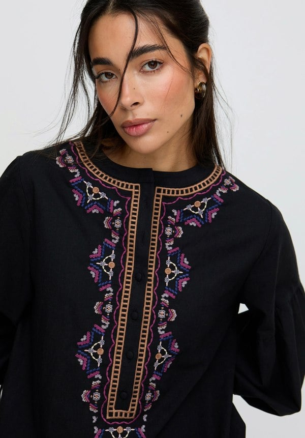 Woman wearing a black outfit with intricate embroidery on a plain background