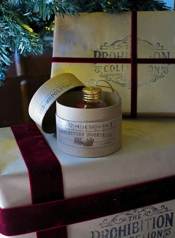 A round cocktail-themed ornament shaped like a ticket sits on top of a wrapped gift box with a gold cap and a red ribbon. A larger matching gift box and green tree branches are visible in the background.