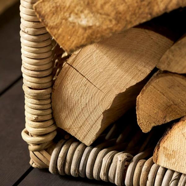 Close-up of the bottom corner of a tall woven Kubu rattan log holder filled with split firewood logs, resting on a dark floor.