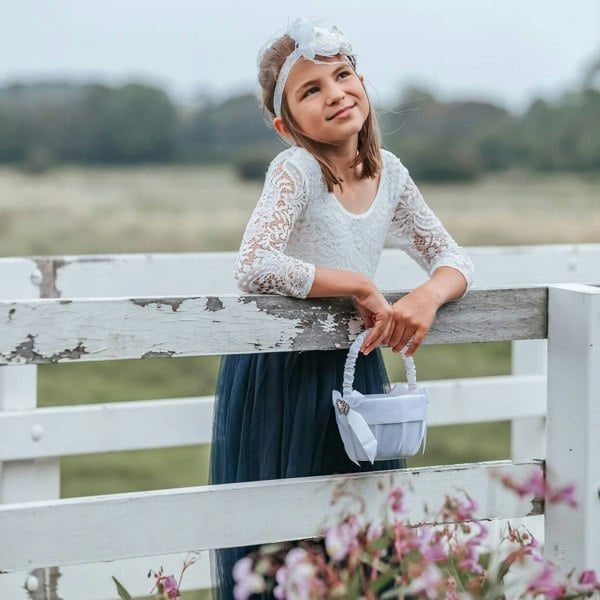 A young girl wearing a navy blue dress with a lace bodice and 3/4 length sleeves, standing by a white fence.