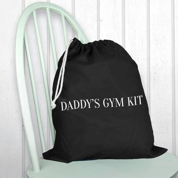 A black cotton drawstring gym bag printed with DADDY'S GYM KIT in a crisp white uppercase font, resting on a light mint green wooden chair.