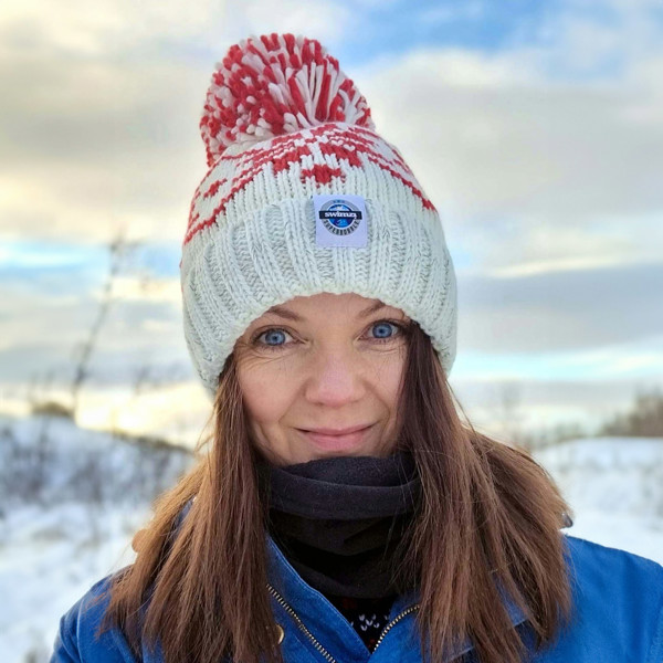 Woman in a tree lined valley is wearing a white Nordic knit bobble hat that has a repeating ruby red large snowflake around the hat and has a big pom-pom and a deep ribbed headband. It contains reflective yarns and is lined in luxury sherpa fleece.