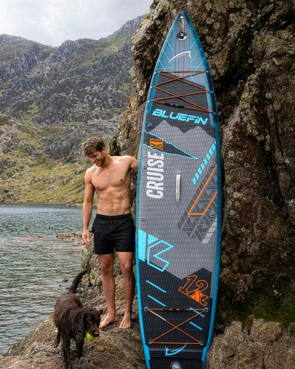 Man standing beside a Bluefin Cruise XL paddleboard on rocky shore, dog nearby, lake and mountains in background.