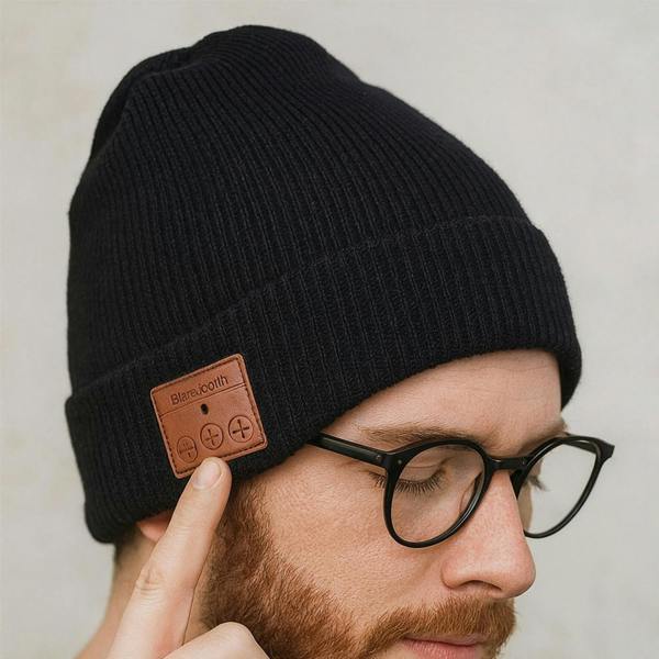 A close-up studio shot of a man wearing a sleek black ribbed wireless beanie hat. He is wearing glasses and touching the leather control panel on the side of the hat, which features buttons for volume and music control.