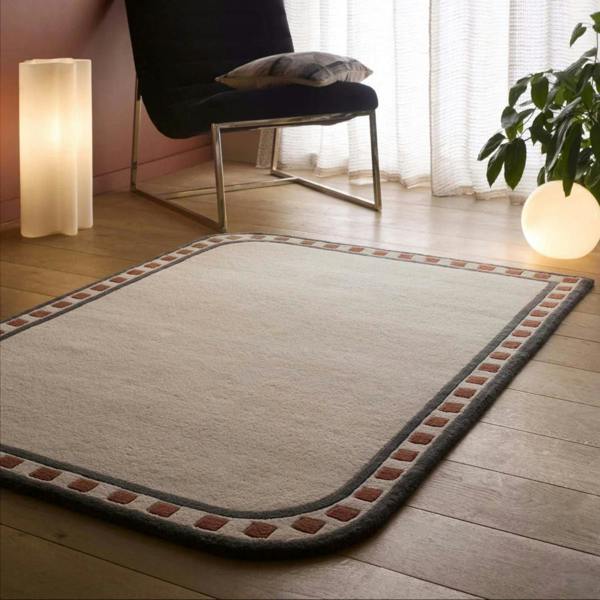 Beige rug with a decorative border on a wooden floor, chair, and lamp in the background.