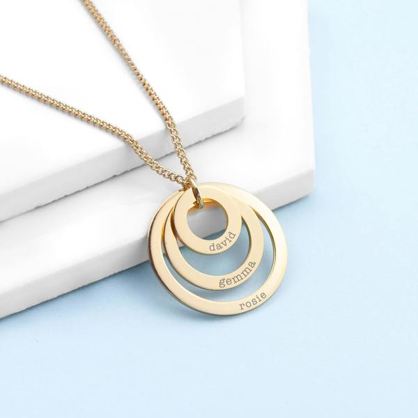 A three-tier gold nesting family ring pendant necklace engraved with the names 'david', 'gemma', and 'rosie' in lowercase serif typography, delicately suspended from a matching gold chain on a tiered white display surface over a soft blue background