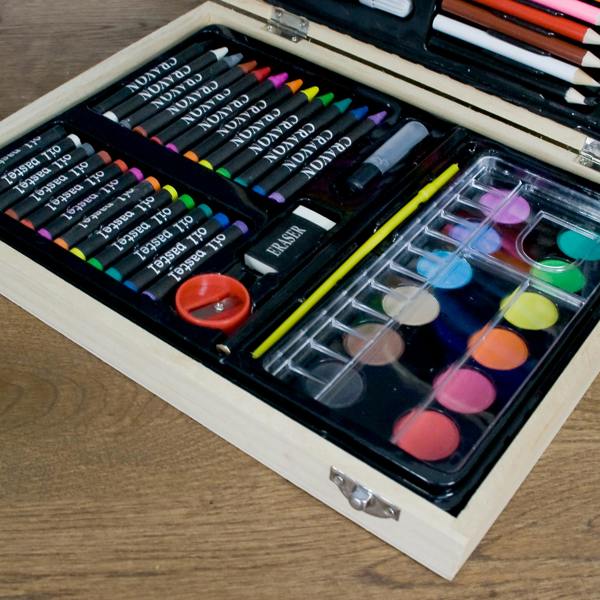 Close-up view of the lower tray in a personalised children's wooden art set, showing oil pastels, crayons, a sharpener, an eraser, and a watercolour paint set with a brush.