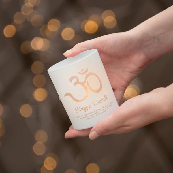A pair of hands gently holding a frosted glass candle holder with a gold Om symbol and a custom Diwali message, against a blurred background of festive fairy lights.
